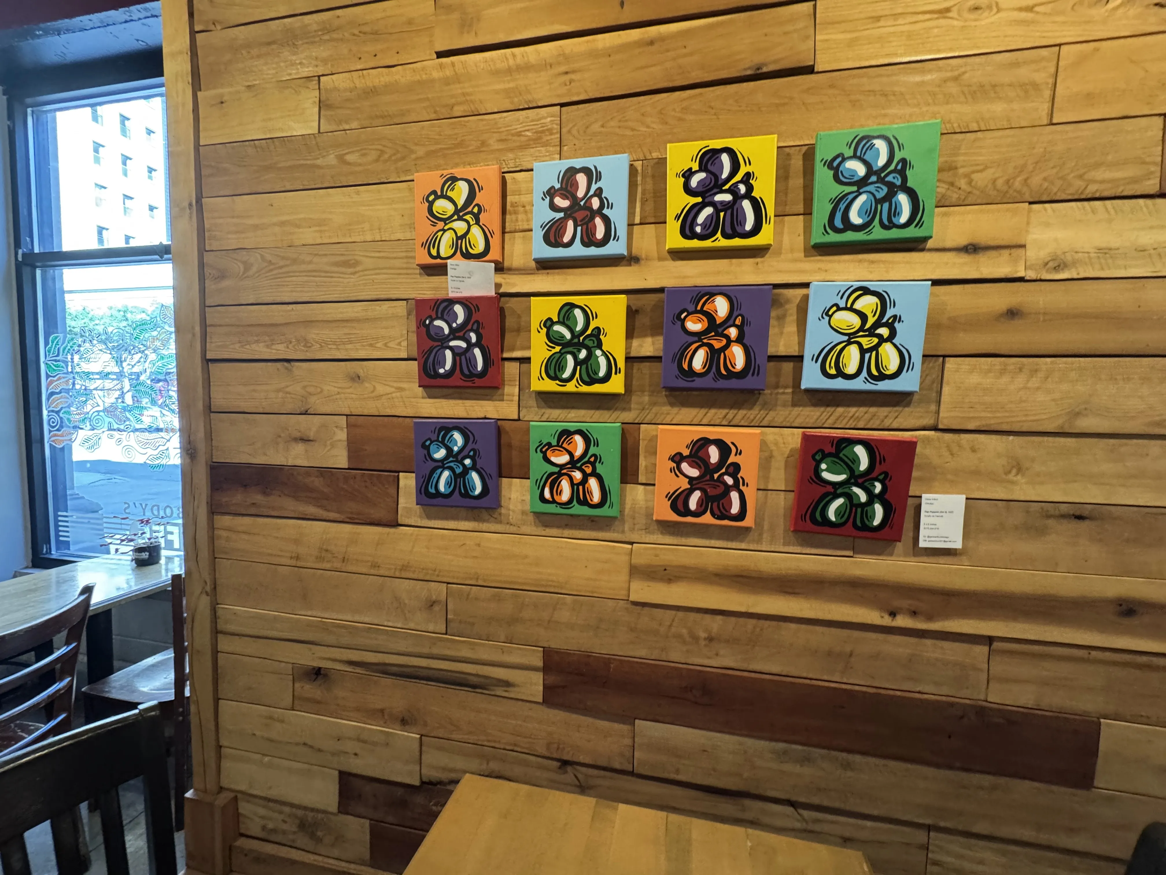 12 small canvases of balloon animal art on a wooden wall above a table
