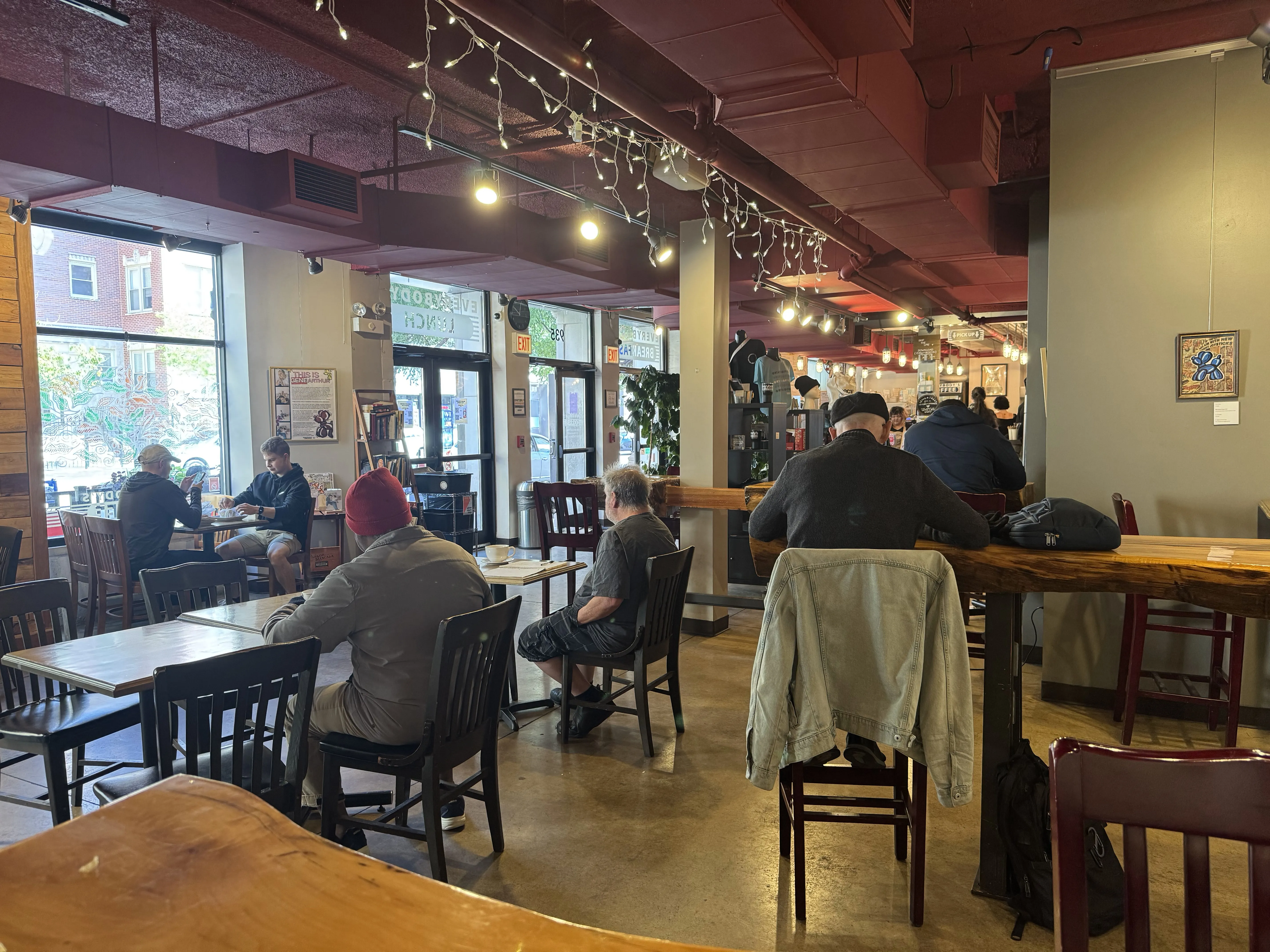 Full view of indoor cafe area, some patrons backs are to the camera. Seated at a high natural-log table, facing forwards towards the front of the cafe. Windows to the left
