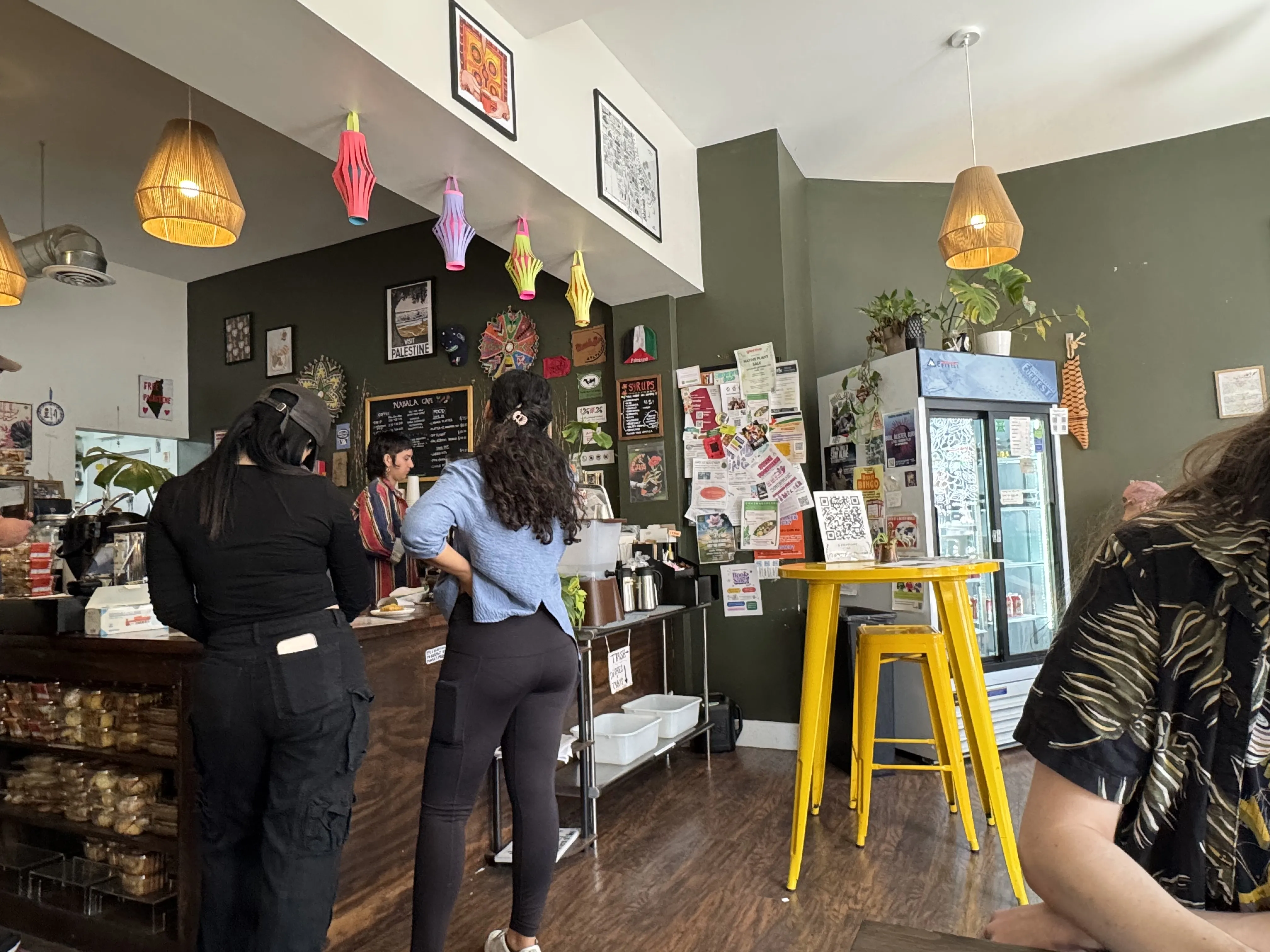 Front of the cafe, some patrons backs facing the camera. Yellow high table has some community fliers/info on it, and the barista counter is visible in the back.
