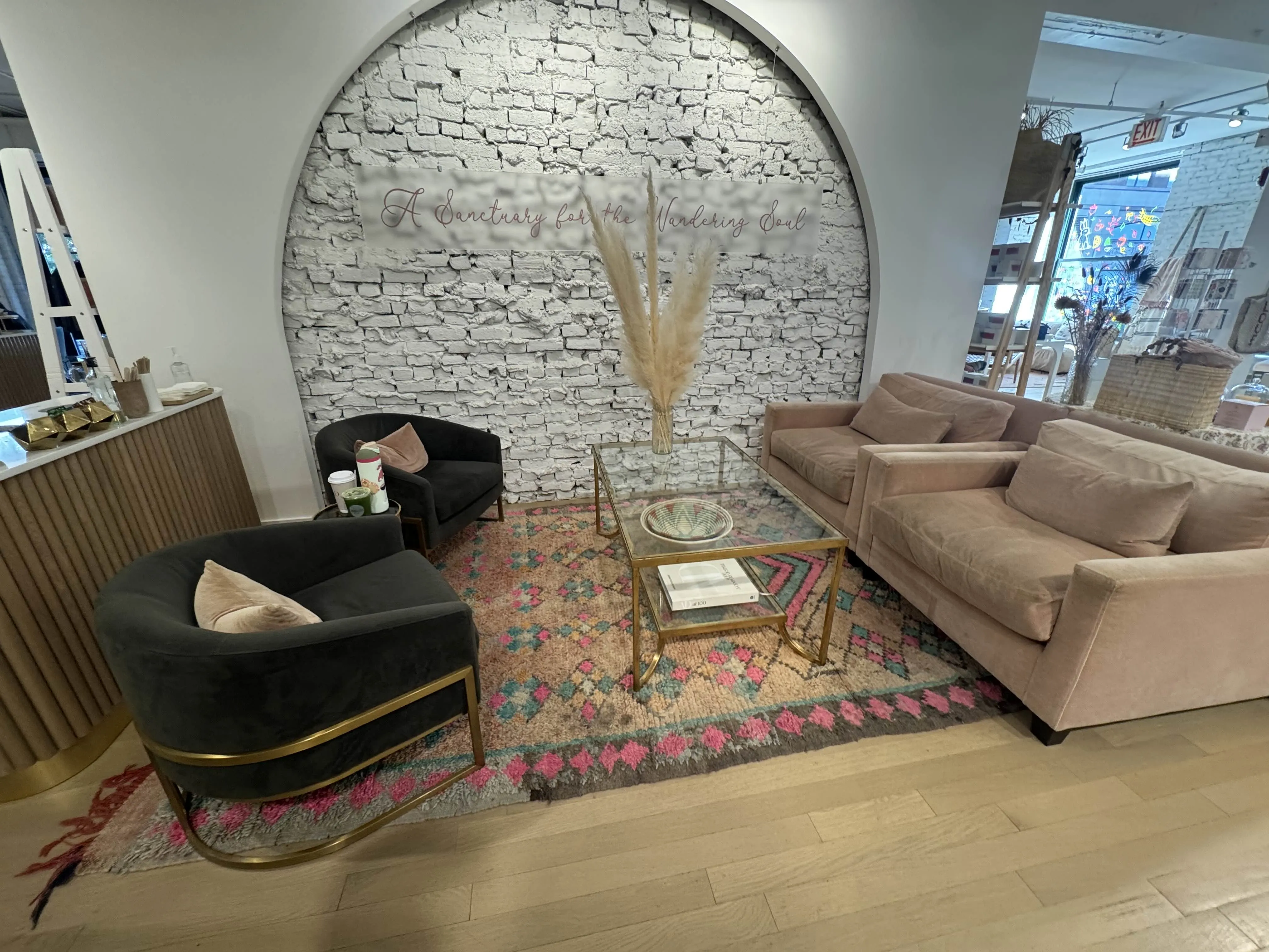 Sitting area right before coffee bar. Two large peach armchairs, a glass coffee table, and two deep green/grey armchairs facing it. The wall behind is a smooth archway made of drywall, displaying the exposed white-painted brick behind. A sign in the middle reads "A sanctuary for the Wandering Soul"