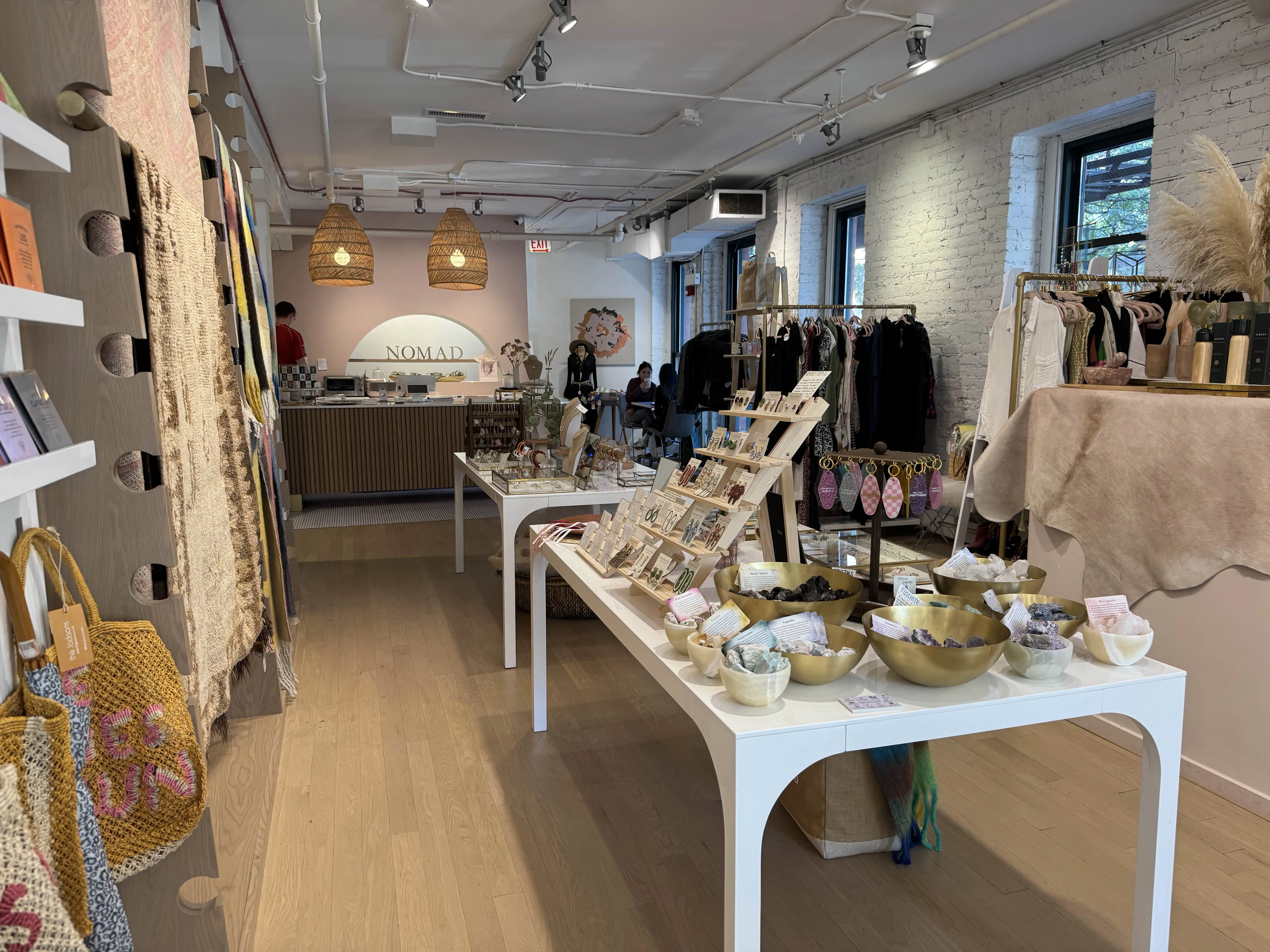 Side of store, the most densely-packed with boutique items. Purses, cards, and rugs on the left. Tables in the center with earings and keychains and fancy rocks. Clothing to the right. In the back the coffee bar is visible with the word NOMAD on a shelf.