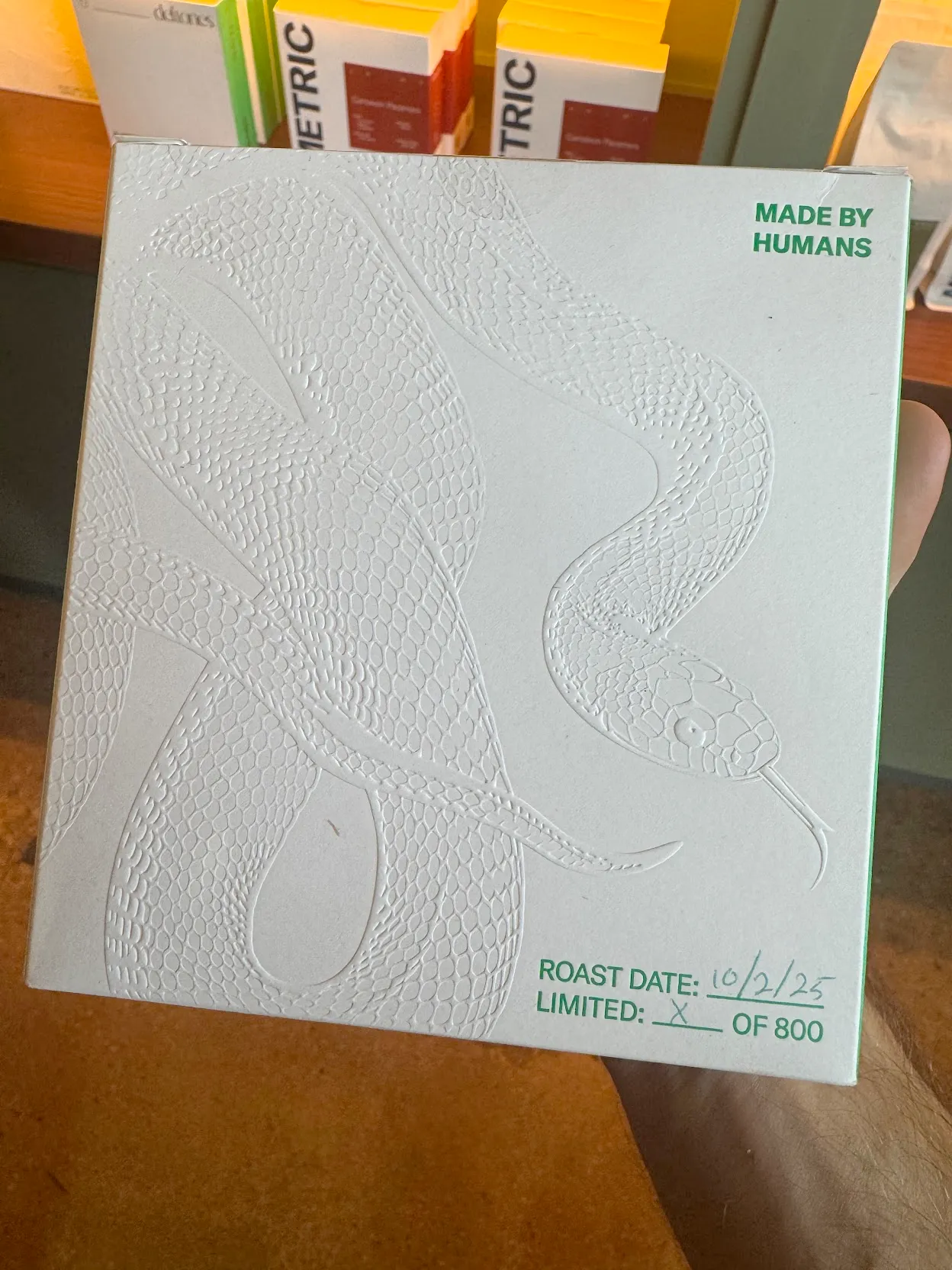 Closeup of white coffee box with embossed emerald tree boa. The writing says "Made By Humans" and lists a roast date and counter of which box of 800 this is