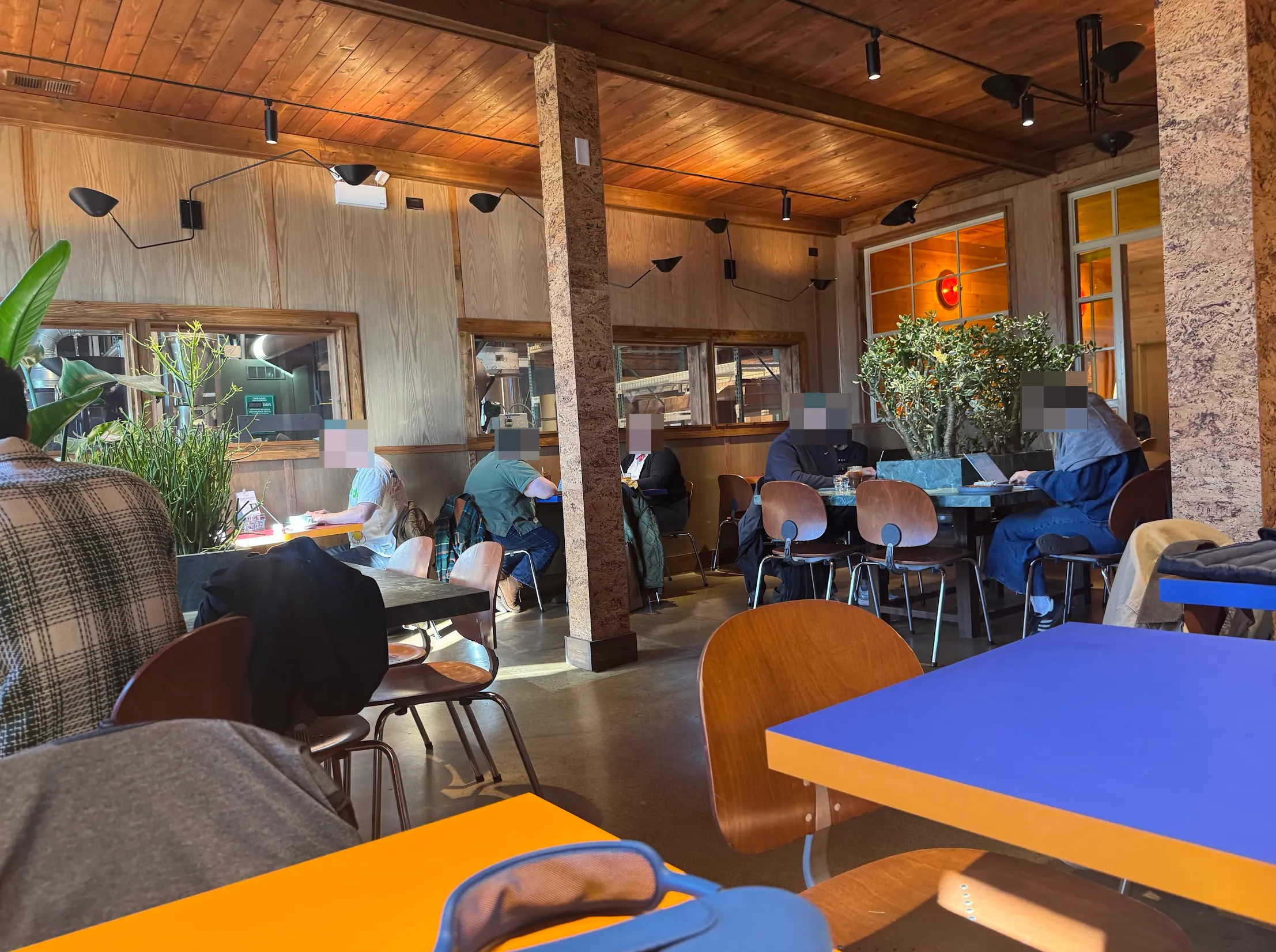 Wide shot of the main cafe area. From the top left, wooden boards and ceiling panels. Below are small windows into the roastery, sealed off from the cafe space. People will blurred faces are seated at 3 varieties of seating, all with treated wooden and metal chairs. In the foreground there are saturated blue & orange cafe tables