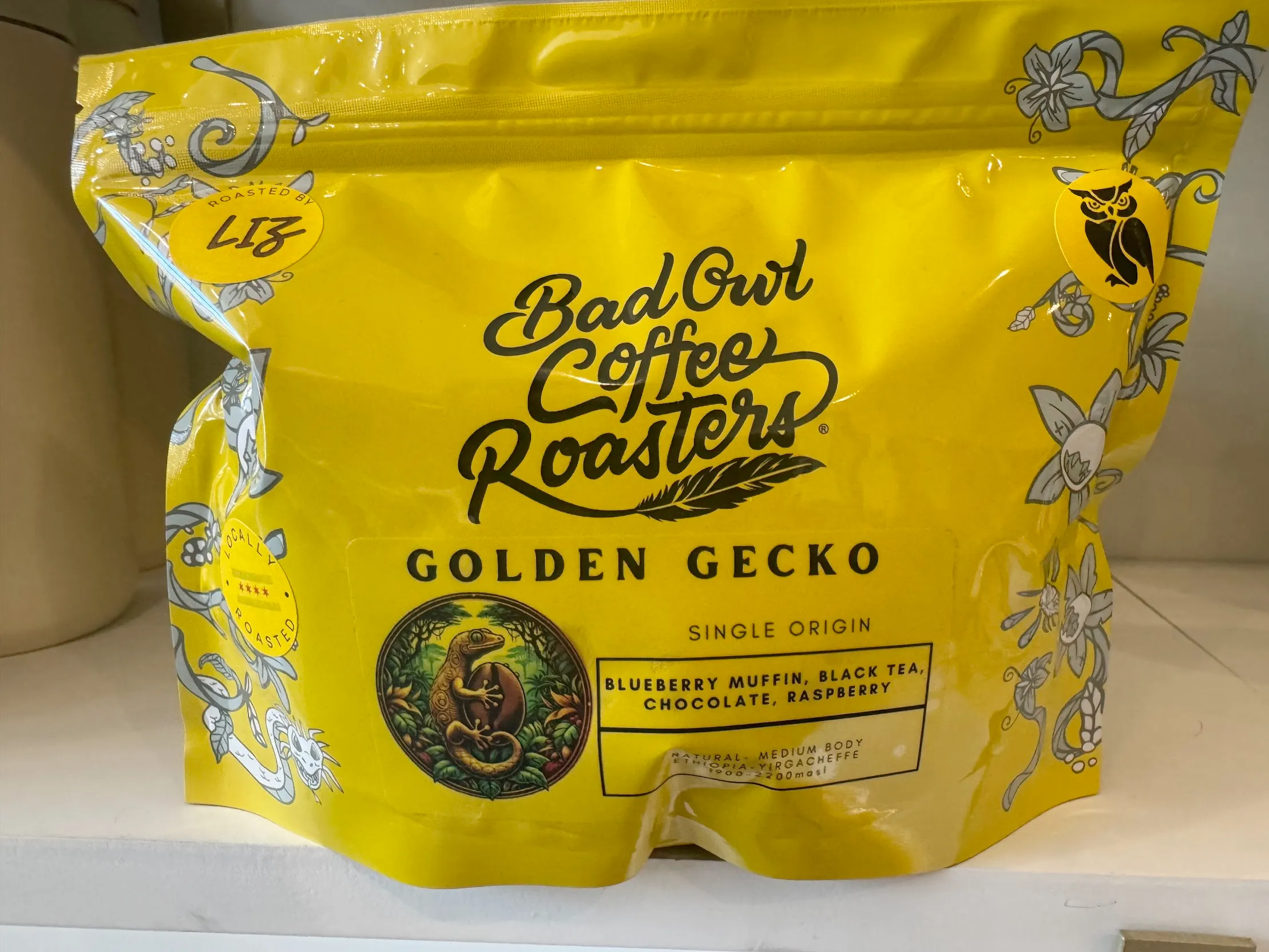 Yellow coffee bag with intricate coffee foliage designs around it, and a sticker on the bottom-left that looks like AI-art.