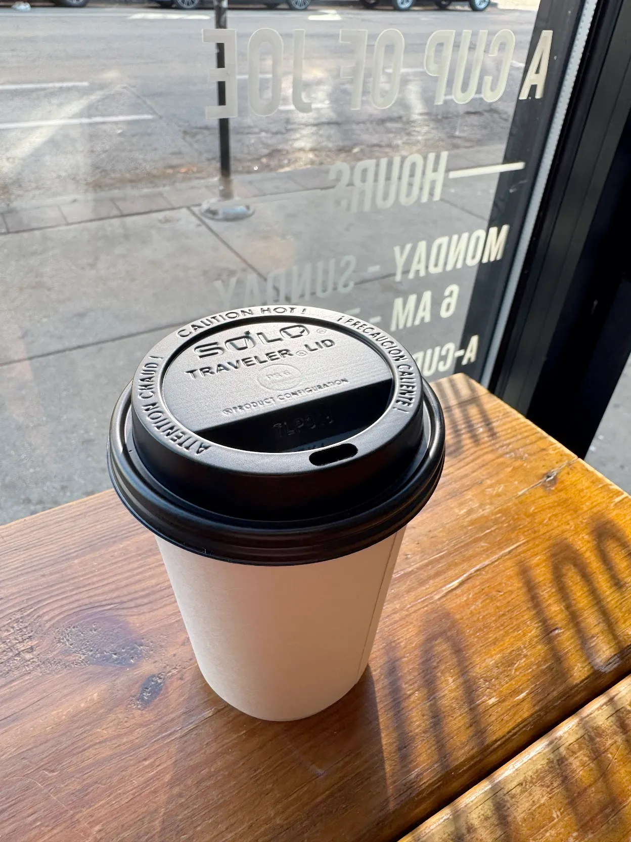 A white travel cup of coffee with black lid on a wooden counter in front of a window at Cup of Joe Pilsen