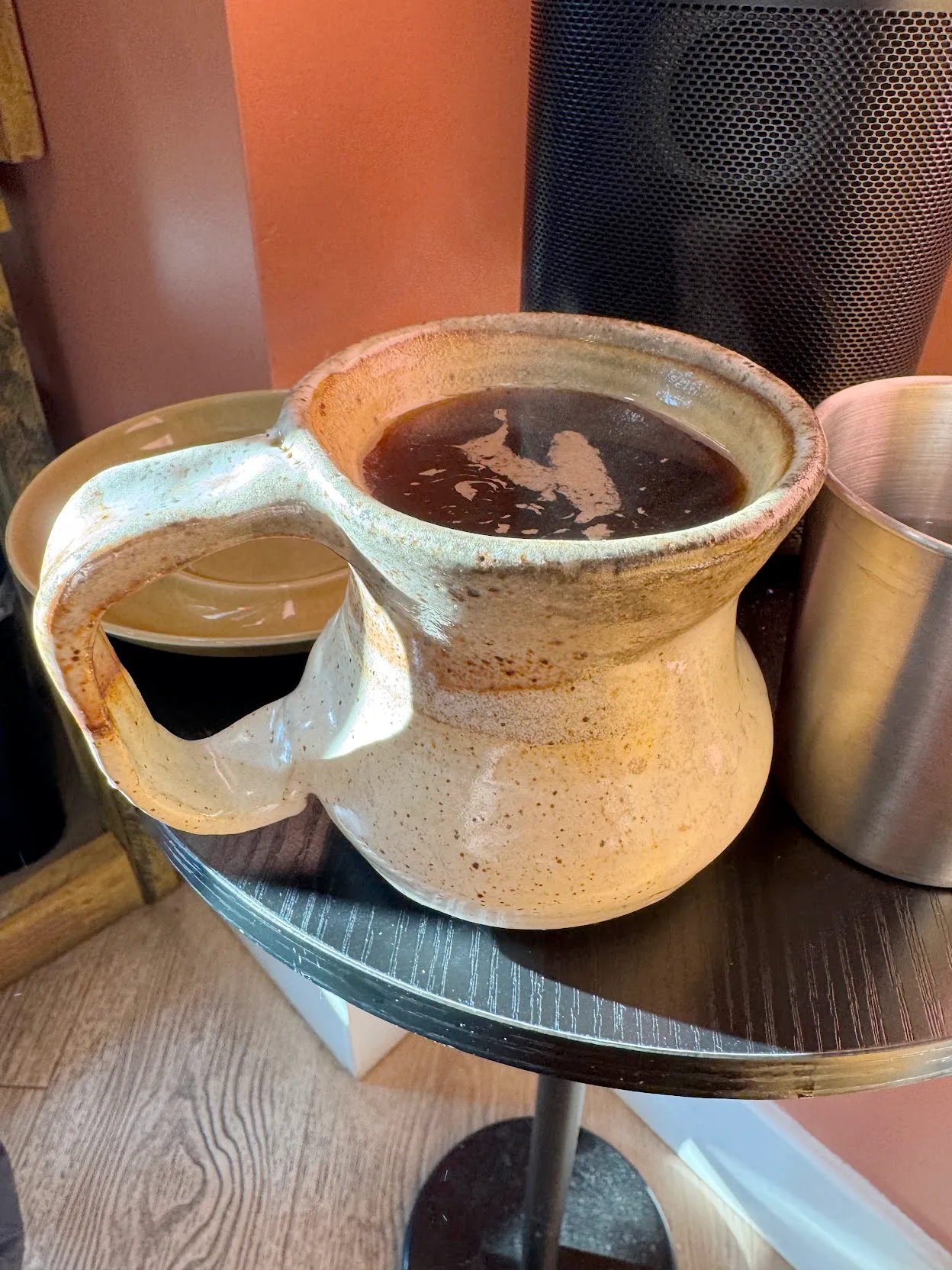 A hand-made ceramic mug filled to the brim with coffee on a small table beside a bluetooth speaker