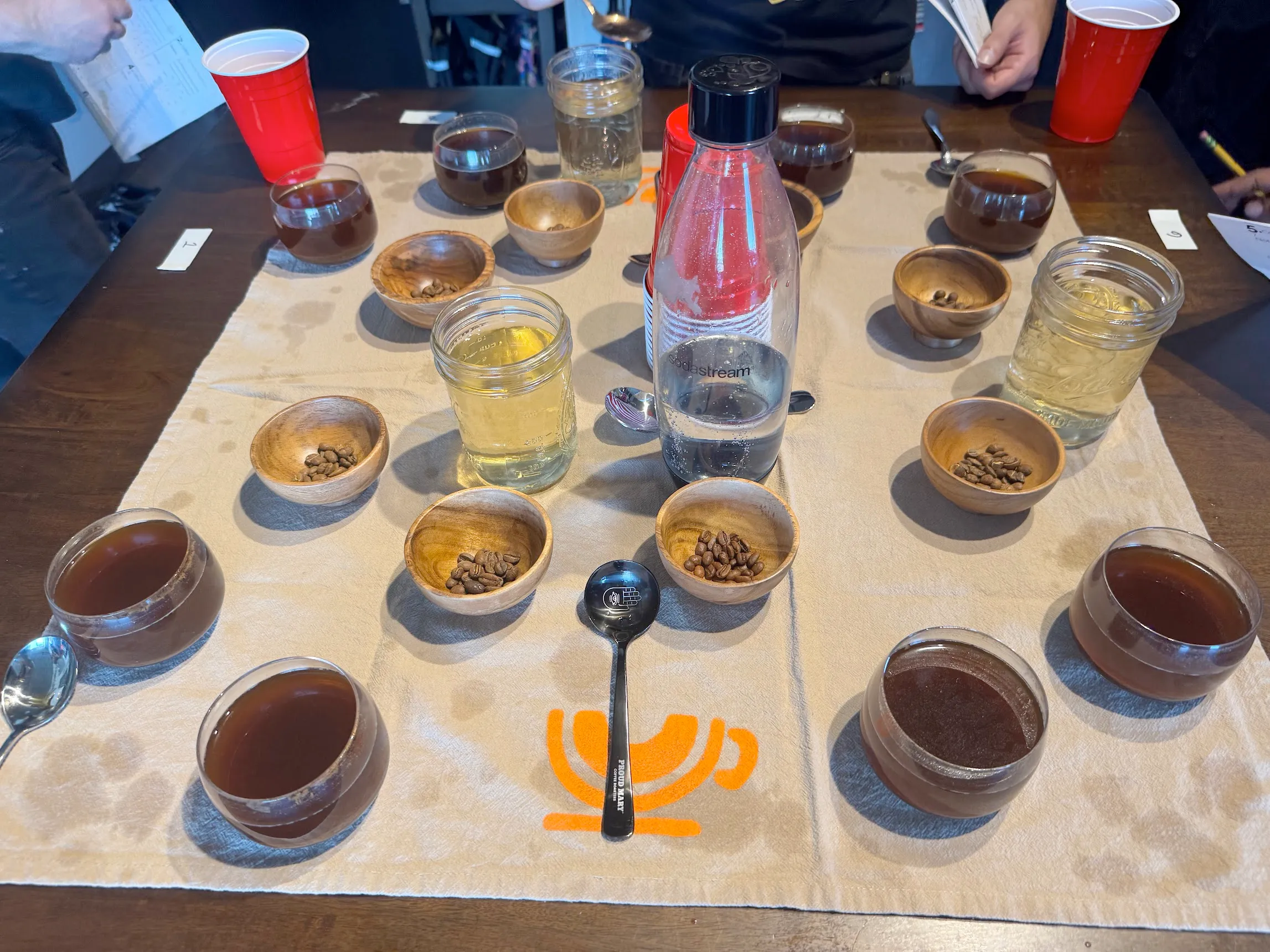 A coffee cupping. On top of a custom tea towel there are 8 sets of: shallow glass bowl full of liquid coffee, shallow wooden bowl with whole coffee beans. There are a few soup/cupping spoons scattered around. There is a large bottle of carbonated water in the center, by some red solo cups. This is all atop a wooden kitchen table. There are some hands holding papers + pencils in the periphery.
