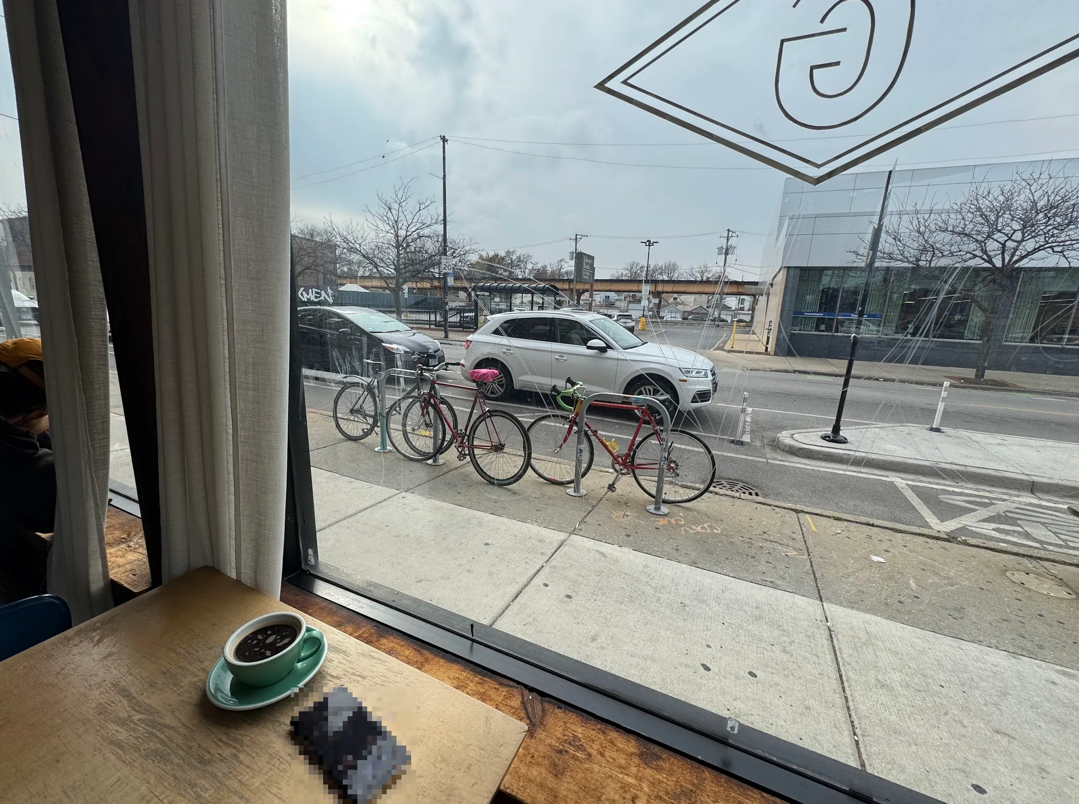 Photo looking out to Milwaukee ave. A coffee mug and phone are on a wooden table in the foreground beside a white-cream colored curtain. The glass has a reversed decal of the Gaslight "G" logo. A car is parked outside and behind some bikes parked on a bike rack