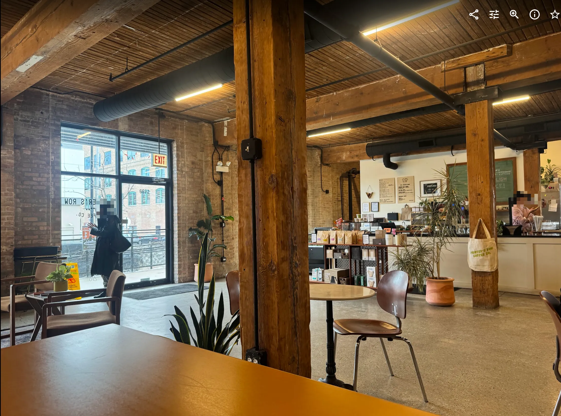 View towards the front door from seating at a large rectangular table. The window to the left lets in the majority of light. There is a lot of open floor space, an empty table, and then the coffee counter at the back.