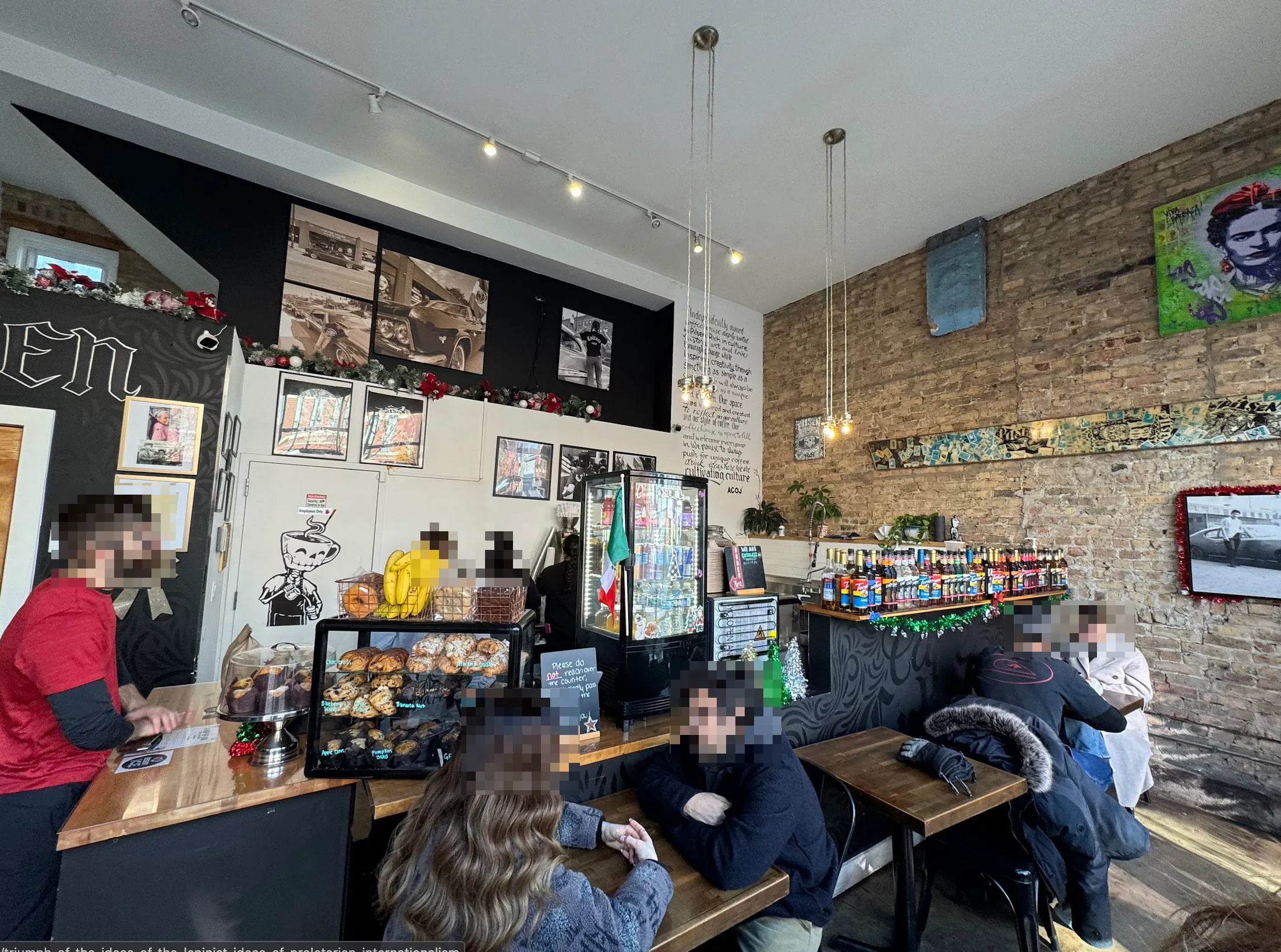 Full shot of the cafe. People are standing ordering coffee on the left, and seated at the sparse cafe seating in the front and right. The wall is covered with Mexican art and photos of old cars