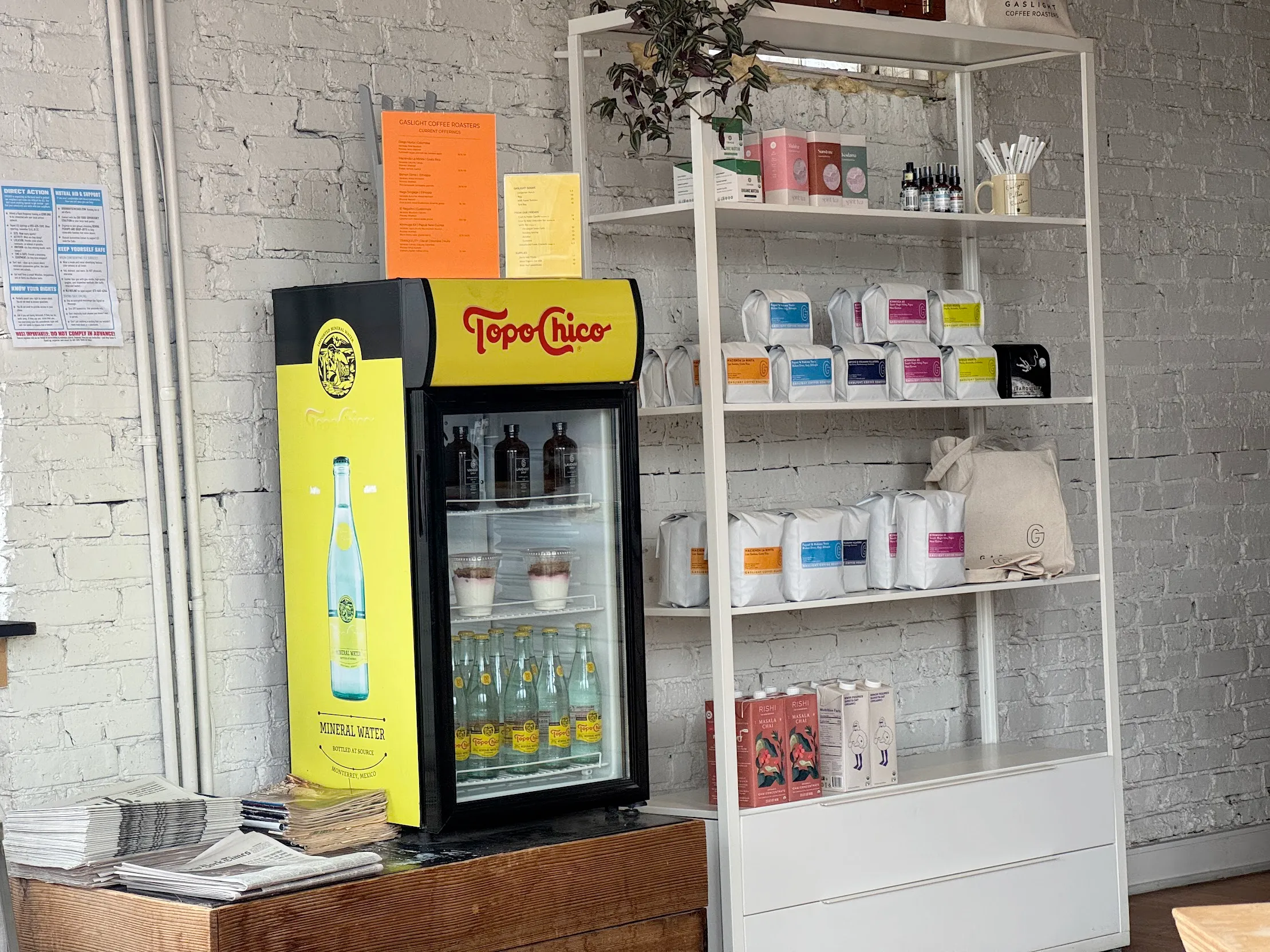 Merch table and small topochico water refridgerator. The shelves are stocked with Gaslight Coffee Roaster coffee and a few pieces of merch.