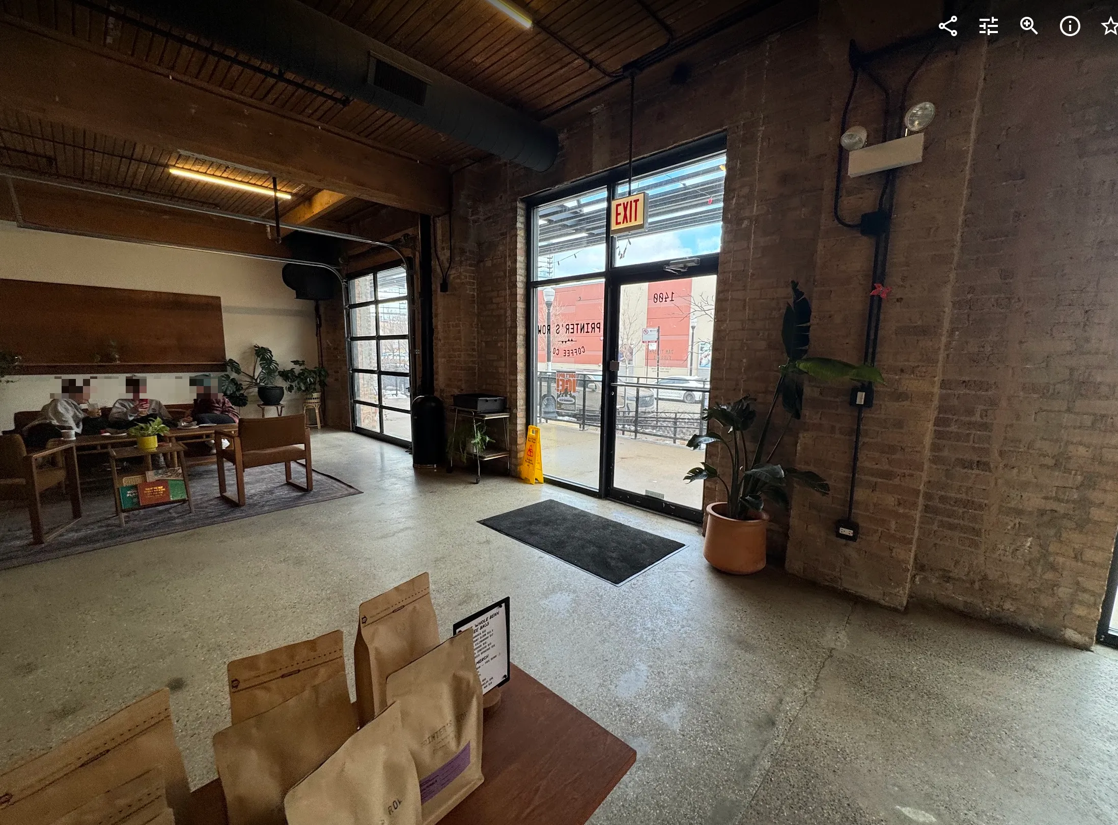 Angled photo of the front door from the register line. The big door + windows on the right letting in light. On the left 3 people sit and talk on the couch. There are coffee beans in the foreground.