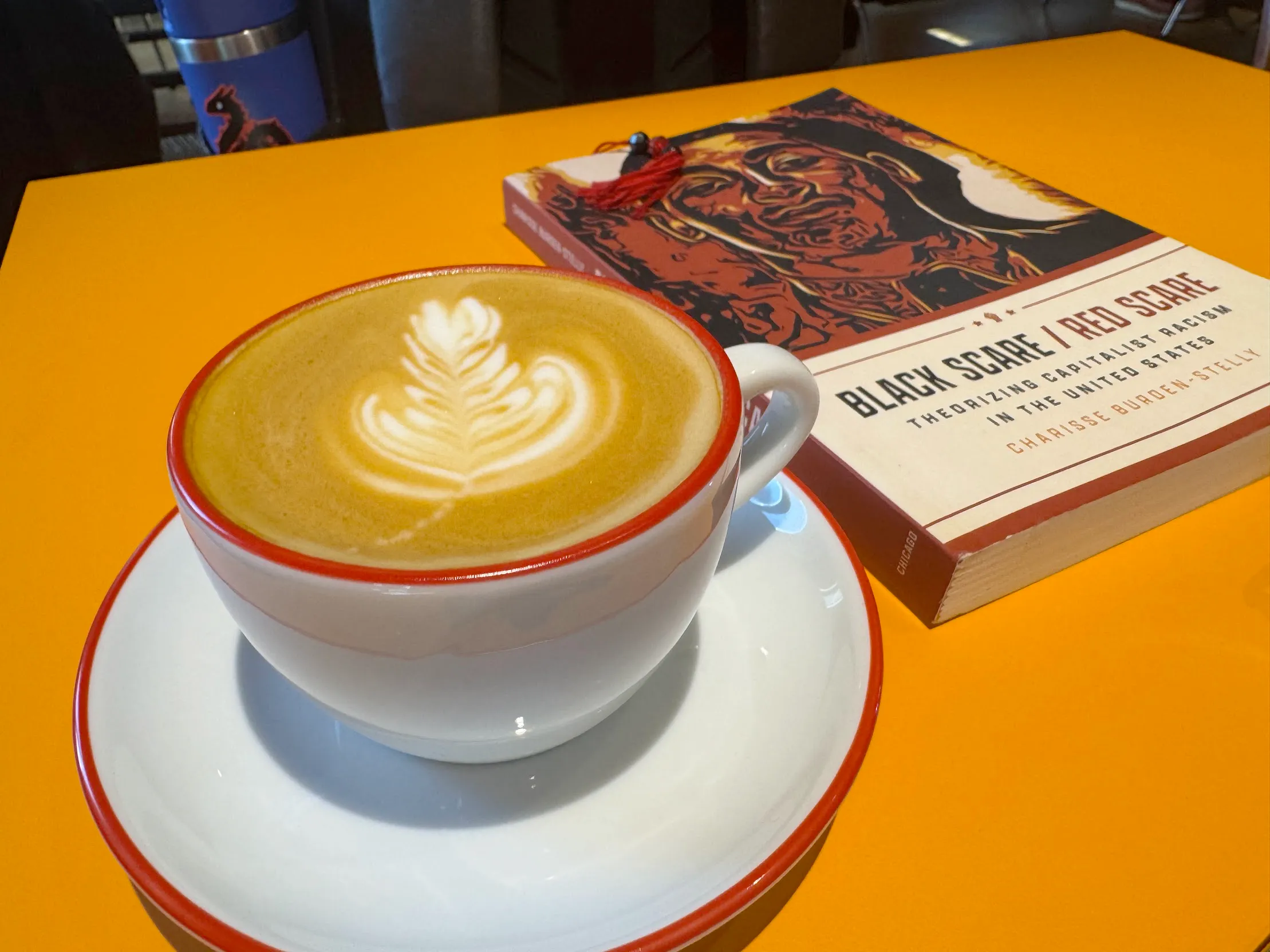 Immaculate latte in a white & red mug, on an orange table, beside a book titled "Black Scare / Red Scare"
