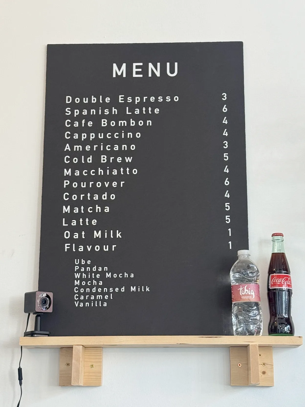 Black and white menu board with a security camera at its base on the shelf. To the bottom right are a bottle of water and glass bottle of coke