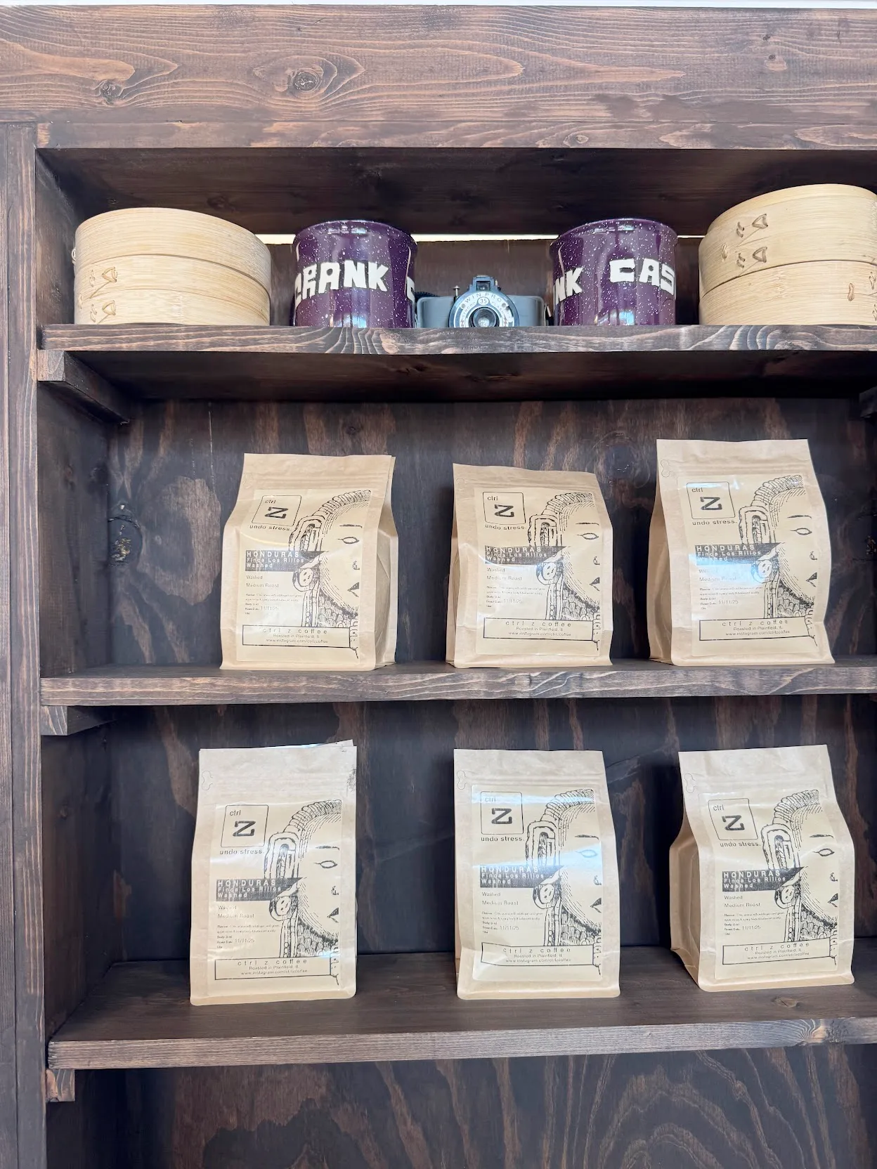 Coffee merch shelf. There are 6 bags of coffee beans on the mid and bottom shelves. The top shelf has 2 handmade purple mugs with steam baskets beside each one. An old school camera is in the center.