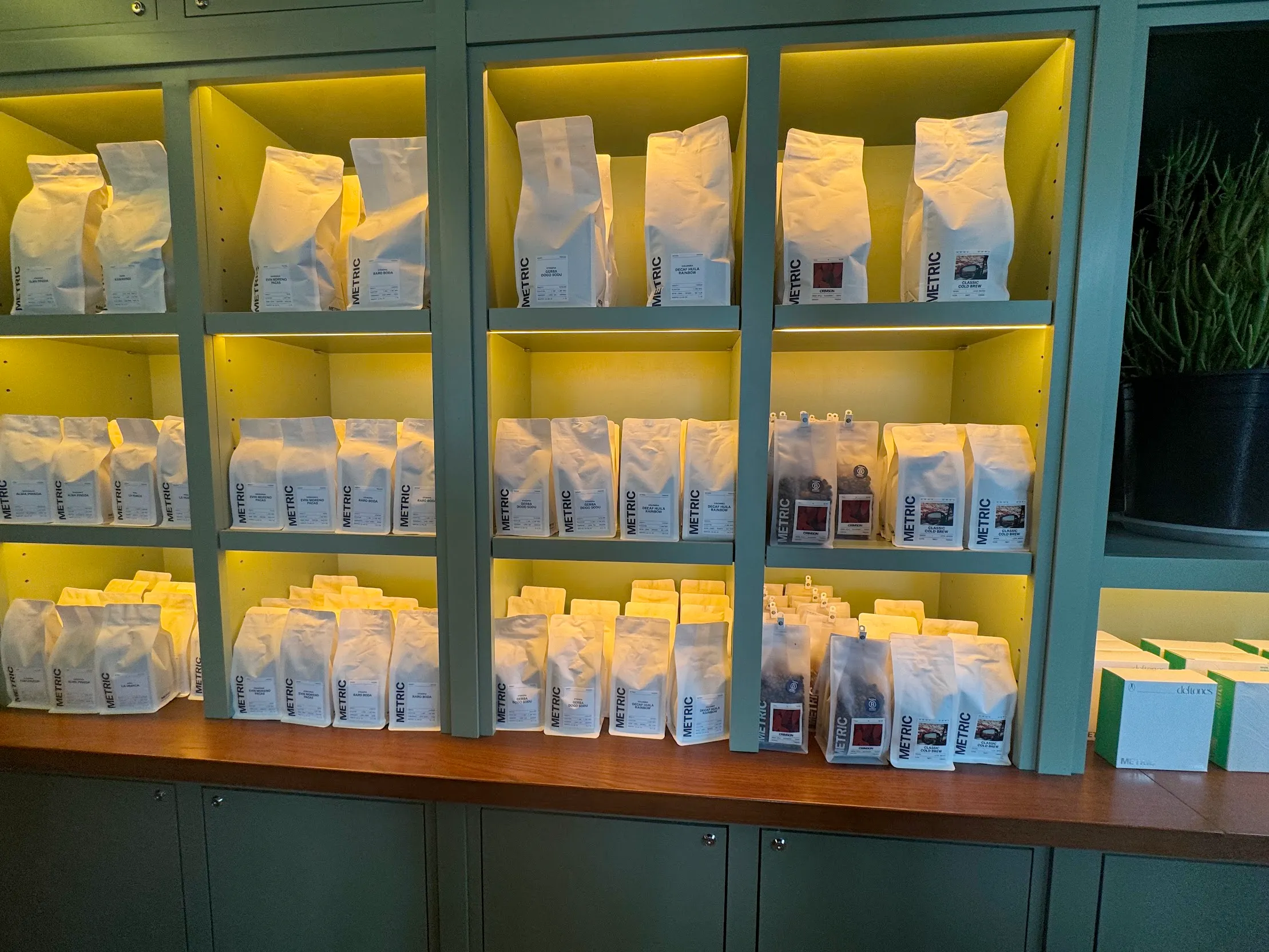 Green and yellow cubbies on the wall full of predominantly white coffee bags. Largest at the top, smaller on the bottom. There's a green cactus barely visible to the right