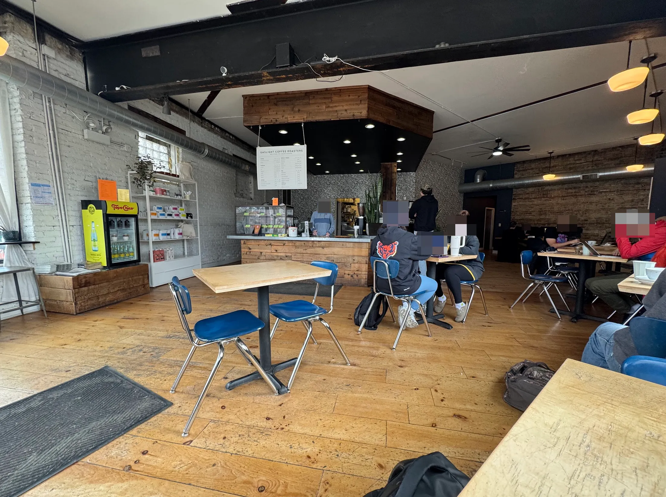Cafe from the front. The main coffee counter and bar is in the center with a menu hanging above a pastry case. Coffee merch and coffee beans for sale are on the left. To the right and throughout are small 2-person cafe tables with some very American public school style blue metal chairs.