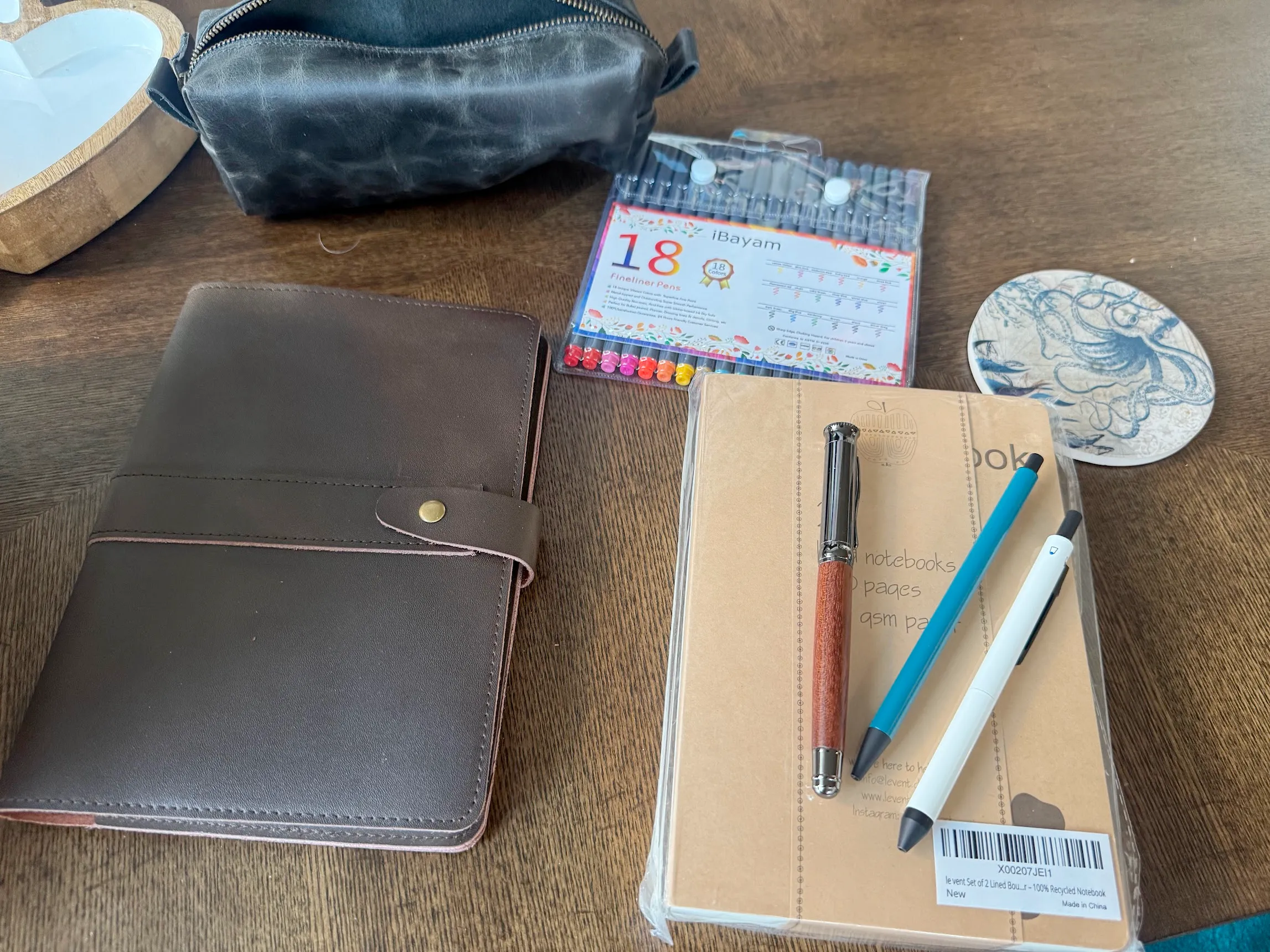 A leather bound notebook on a brown coffee table beside a lighter brown notepad with 3 pens on top of it. Above are 18 colored fine-tip pens beside a leather pencil pouch