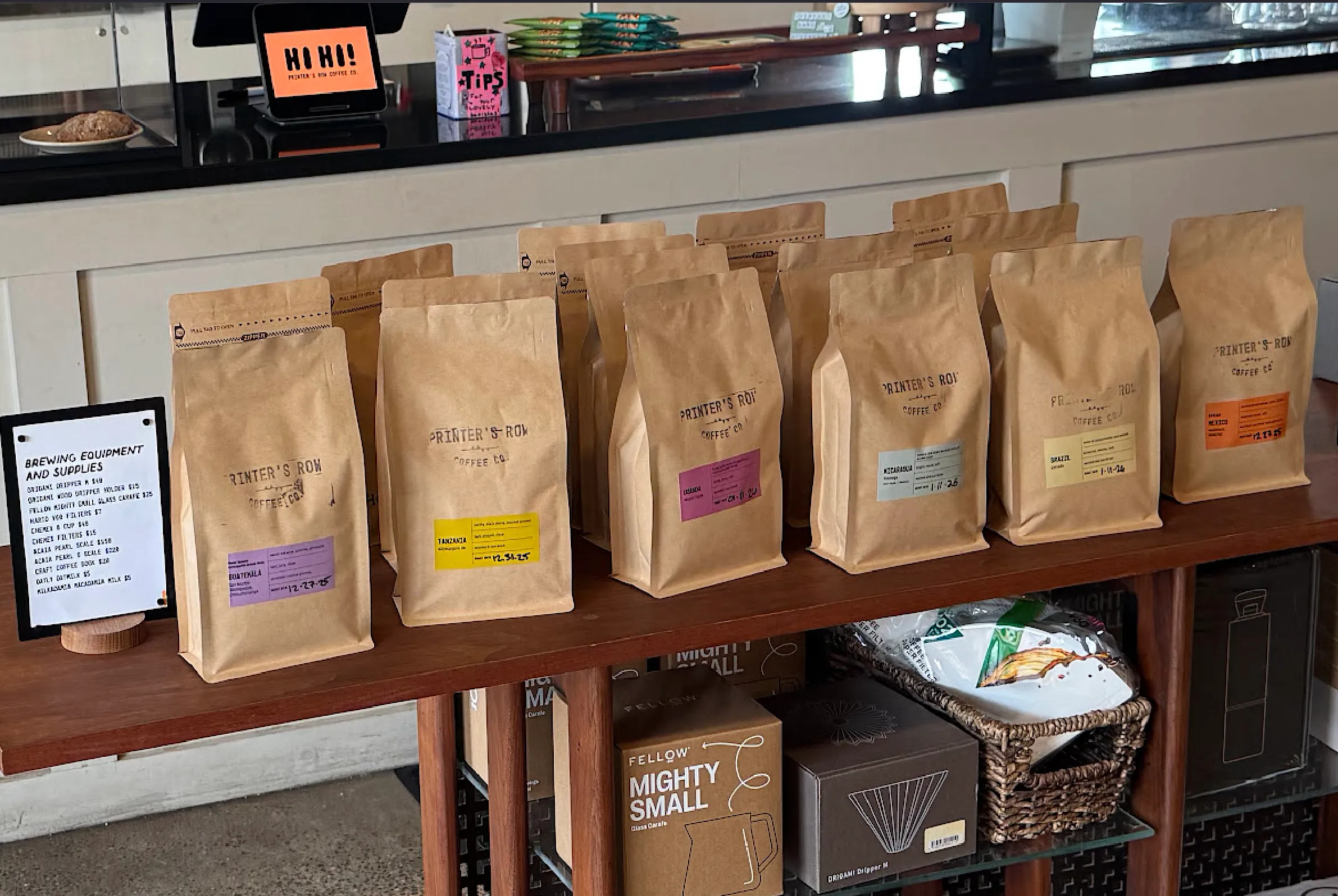 Close-up of coffee bags atop a dark brown wooden stand. The bags are light brown, stamped with "Printer's Row Coffee" and varied colored stickers denoting the specific roasts
