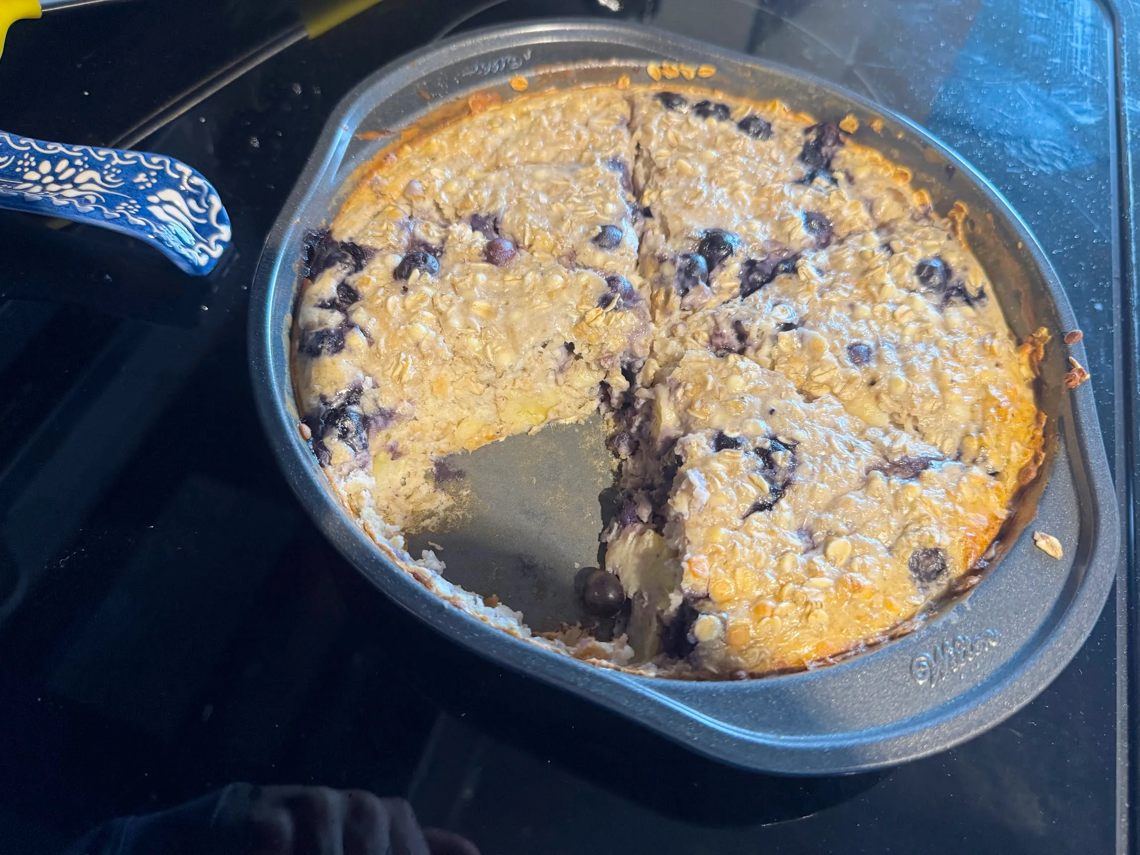Circular oat cake with blueberries and one slice missing inside of a metal baking tin, atop a glass stovetop