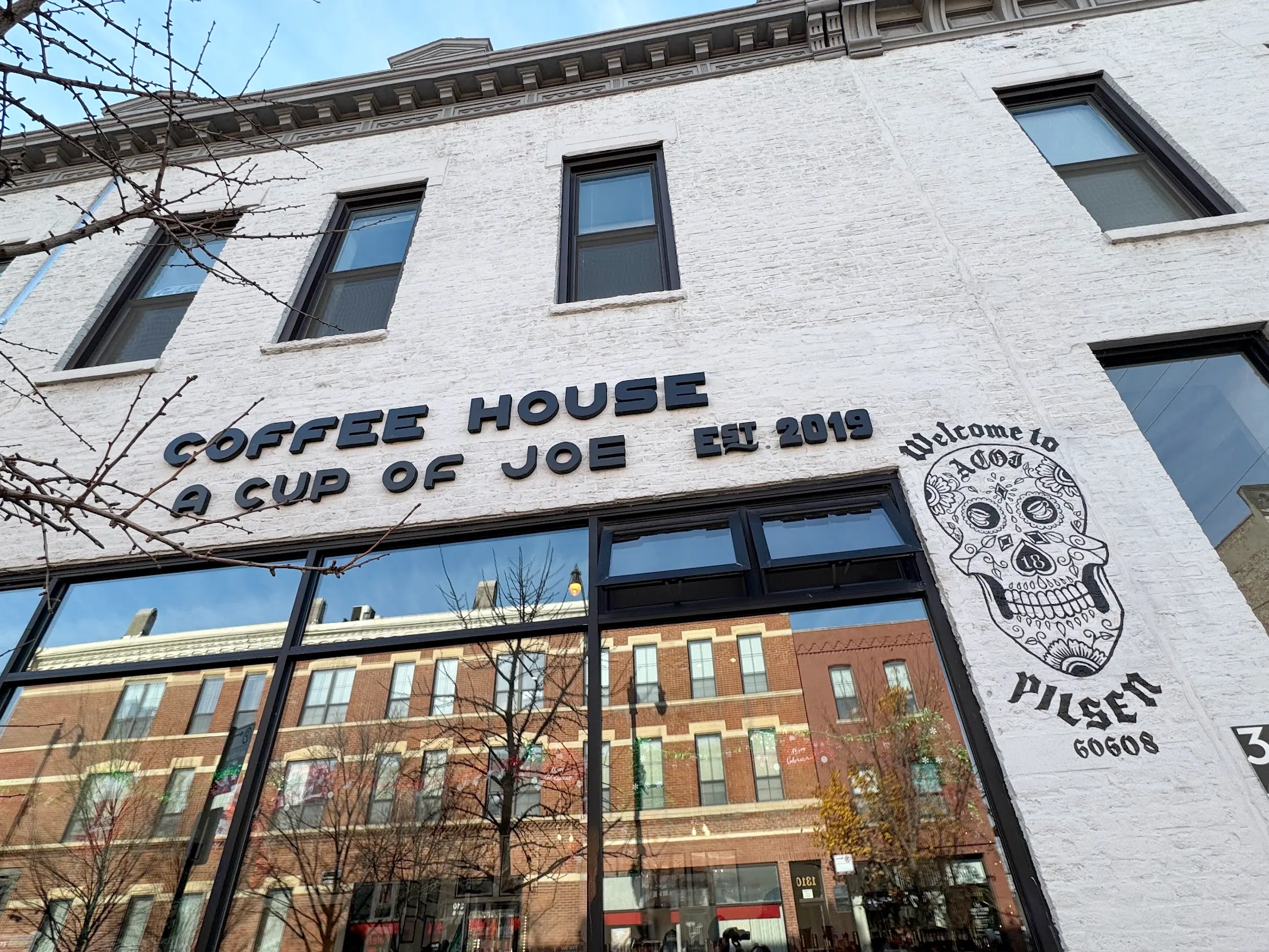 Extreme angle photo of the Cup of Joe coffee house. A white brick building with large reflective glass underneath the store name. On the right side is a Mexican candy skull decal that says "Welcome to Pilsen 60608"