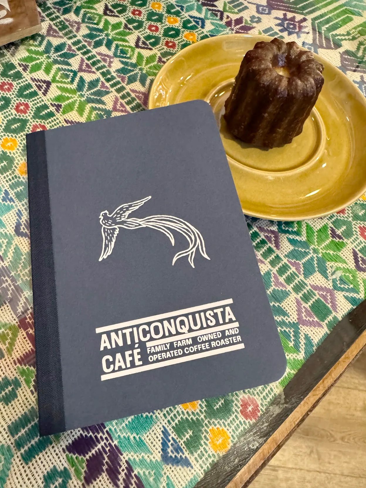 A blue notebook labeled Anticonquista Cafe Family Farm Owned and Operated Coffee Roaster atop a green and blue woven mat beside a yellow plate with a brown cylindrical pastry