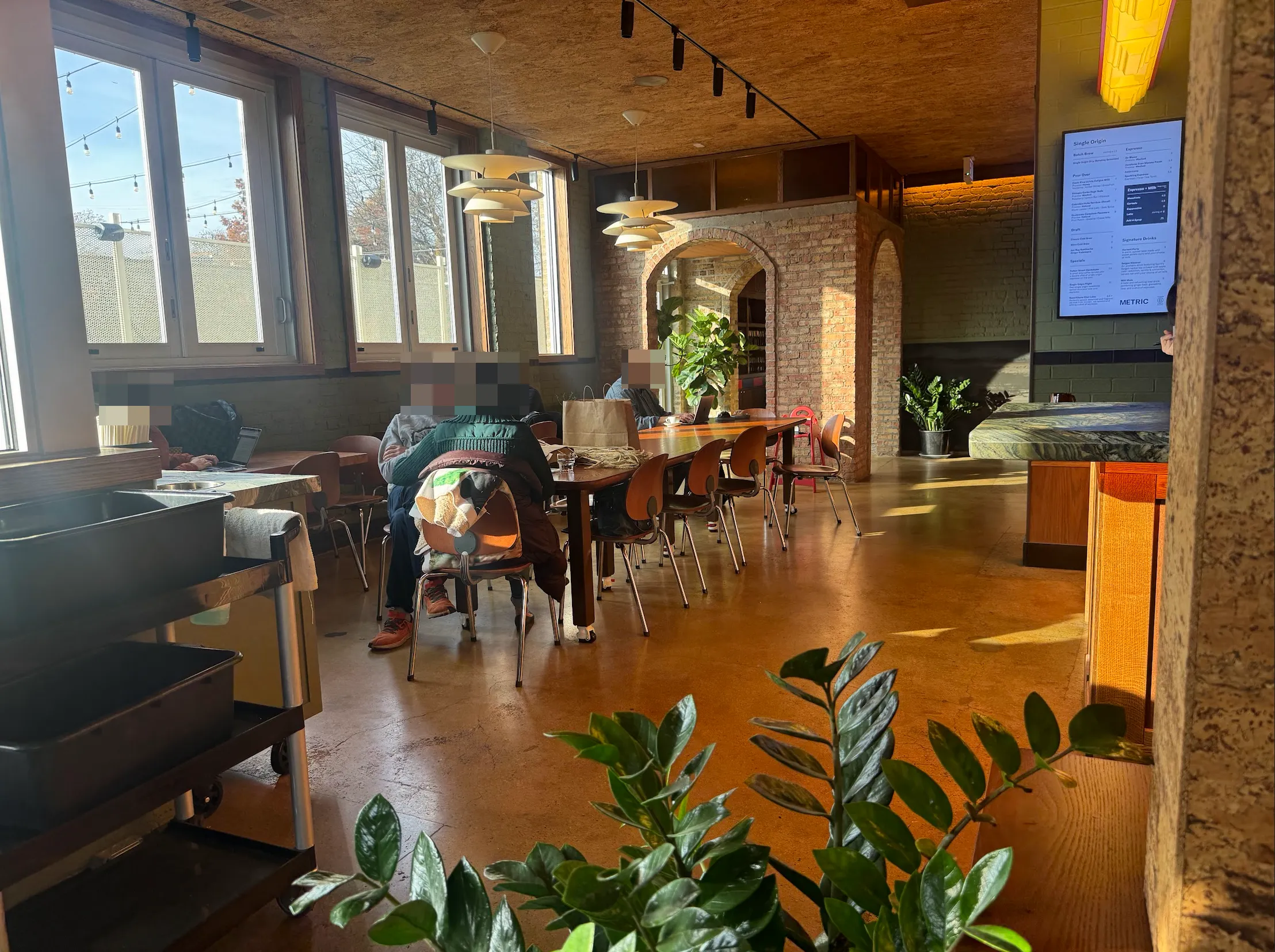 Entryway cafe space. Large windows on the left open into a cafe space with a few small tables and large central "meeting" style table with chairs. There is an archway in the back leading to more of the cafe. There's a bussing station in the foreground on the left. A zz plant. And on the right the barista table is visible.