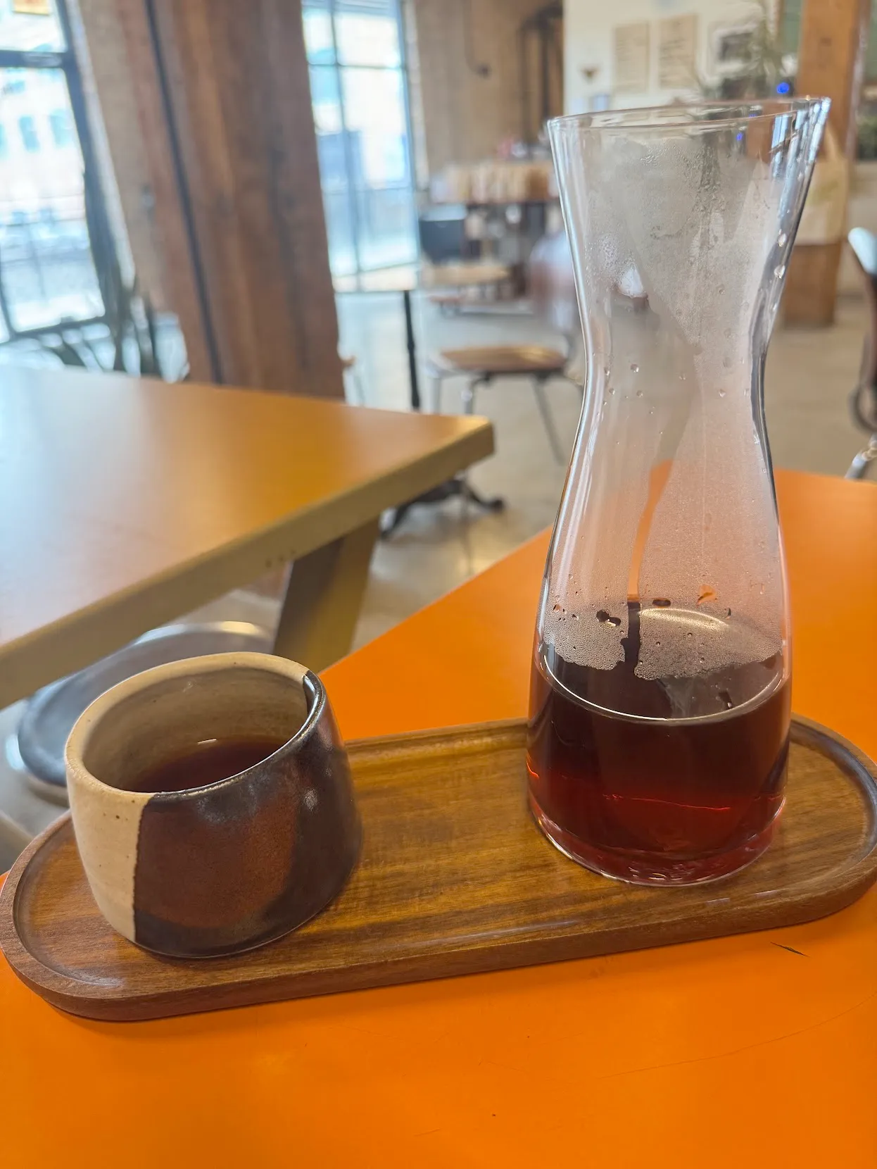 A pour-over served on an oval wooden tray. The cup on the left is a small grey and dark wood cup. On the right is a tall pitcher with a light brown coffee