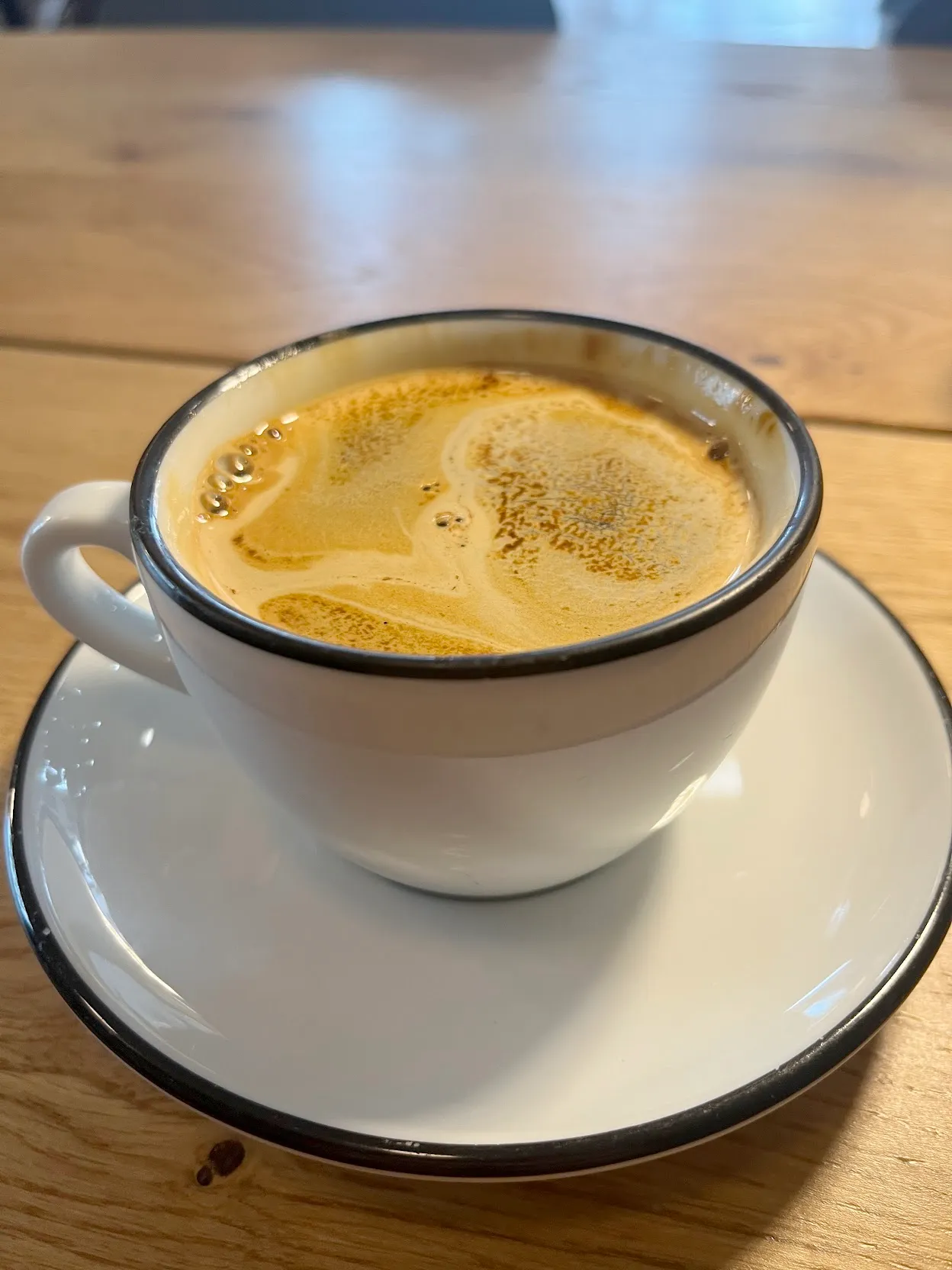 a classic short americano in a white mug with black accents. The espresso is striped and complex. The mug and saucer are sitting on a wooden table