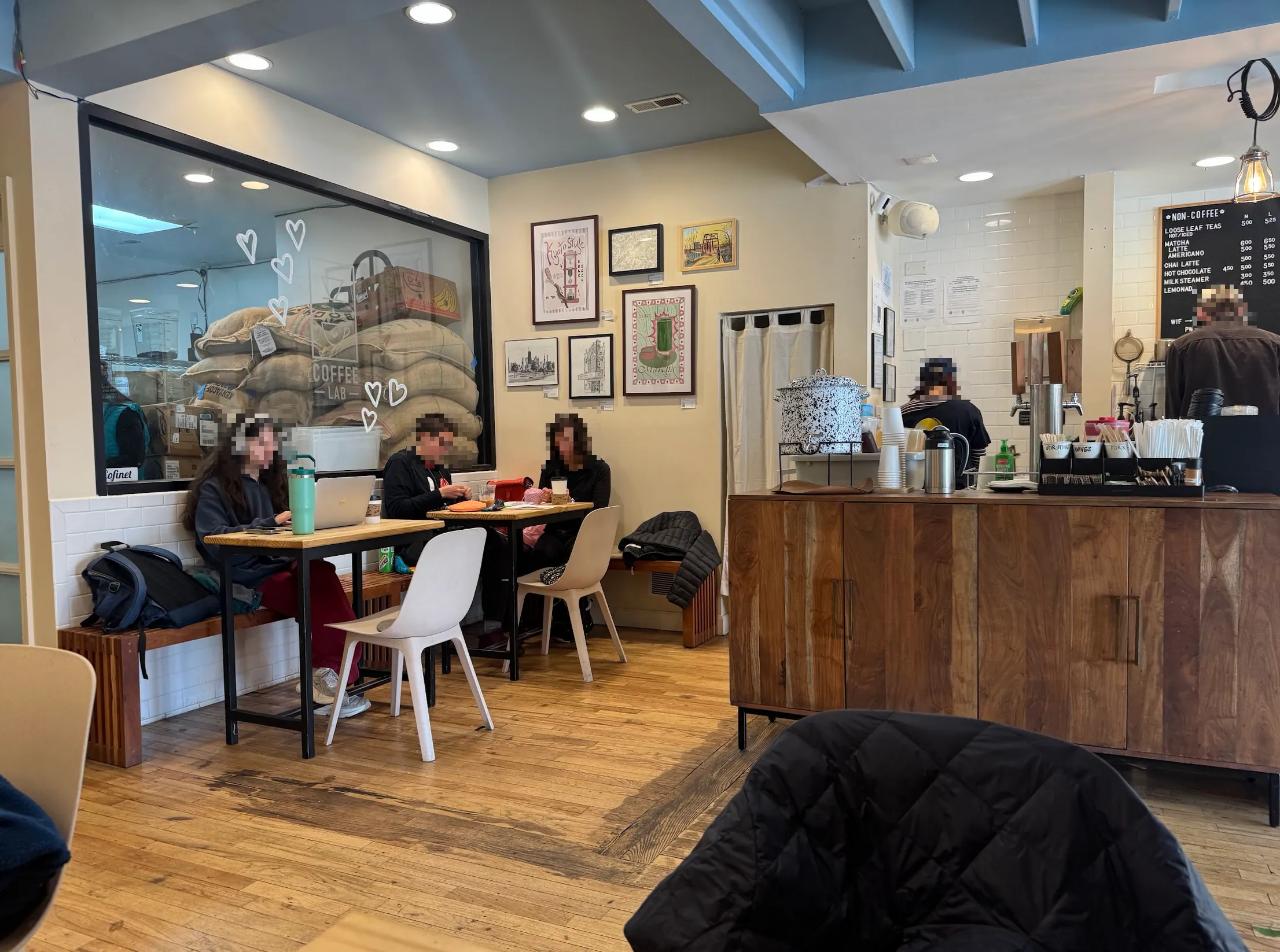 Cafe view. Coffee self-service bar is in front, a warm wooden cabinet with water and sugar etc. Behind it is partially visible coffee bar (espresso machine and syrups). To the left are 2 of the 5 total tables in this cafe. People are sitting at them with laptops out, faces blurred.