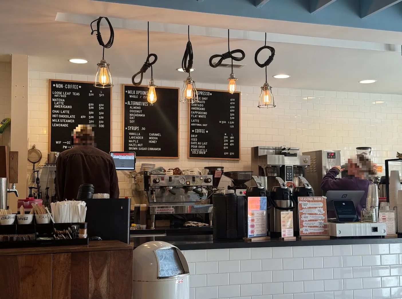 Full coffee bar view. Two baristas with blurred faces stand in front of coffee equipment. Peg letter boards on the wall behind them list the cafe's prices and offerings