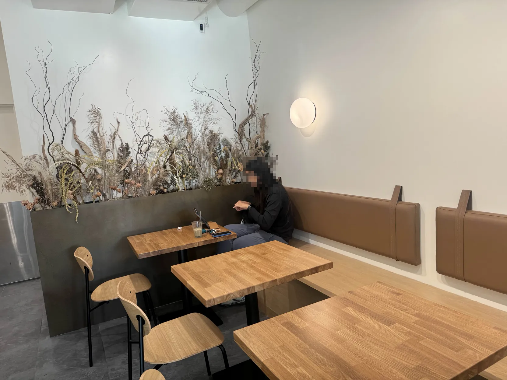 Cafe seating. A drab-toned fake plant collection at the back in a metal rectangular planter. In front, 3 2-person cafe tables with alternating chair or booth seating. A single person sits in the back corner, face blurred