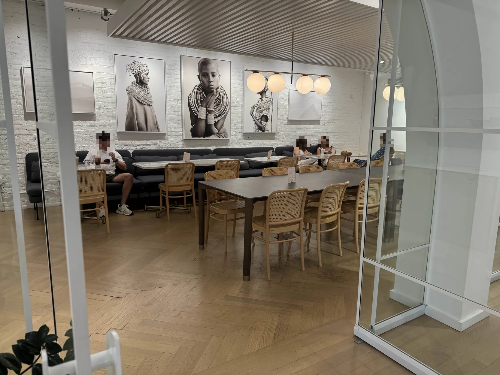 Traditional cafe seating area beyond double-doors featuring many long tables with wicker-backed chairs, and some couch/booth seating. Various photos on the back wall, and people with blurred faces having conversations sitting at various tables