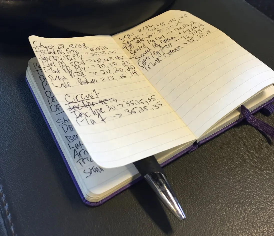 An old picture from 2018 of my purple paper logbook with some exercises and dates written on it. There's a pen wedged between some pages. The notebook is sitting open on a weightlifting bench with 2 plates visible behind it