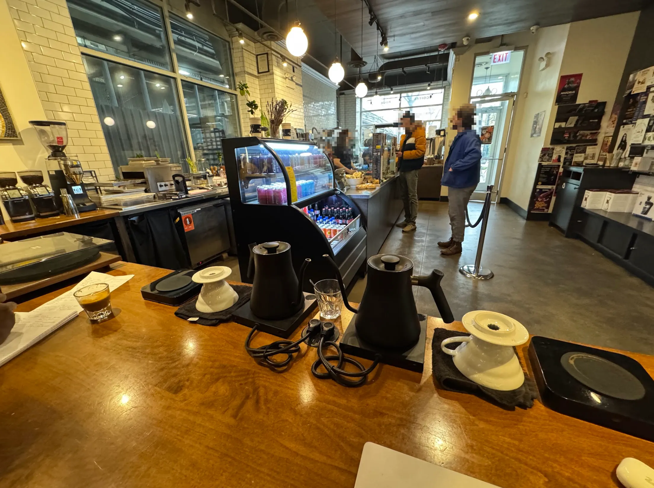 View of cafe entryway. Door and windows to the back, coffee bar and drink fridge to the left. The foreground counter has a kettle.